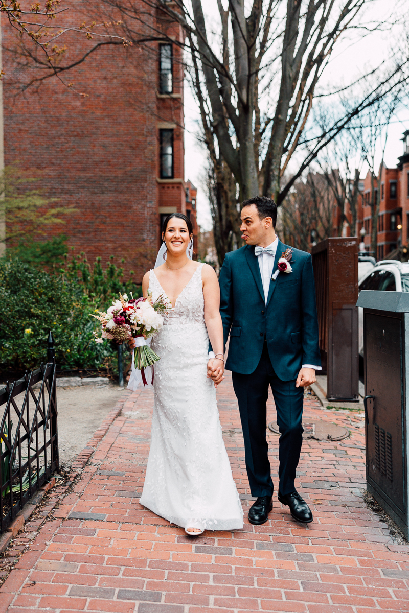 A joyful bride and groom hold hands while walking on a brick pathway, surrounded by blooming greenery and historic buildings.