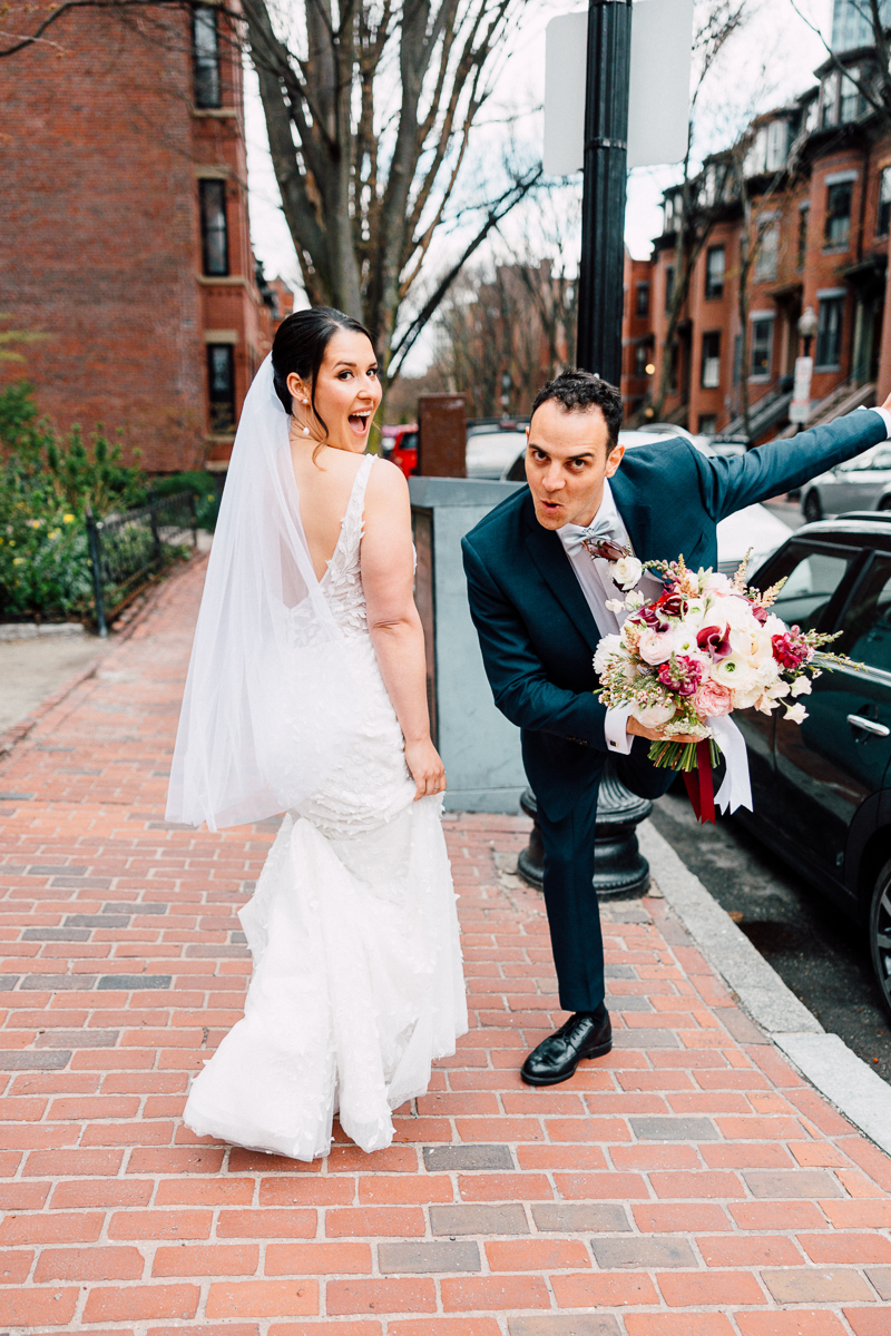 A bride and groom capture a playful moment outside, with the bride smiling and looking back while the groom poses humorously with a bouquet.