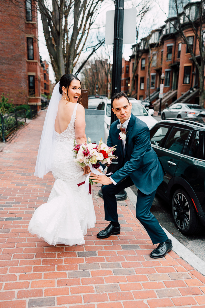 A bride playfully looks back at her groom while he pretends to pull her away, capturing a lighthearted moment on a city street.