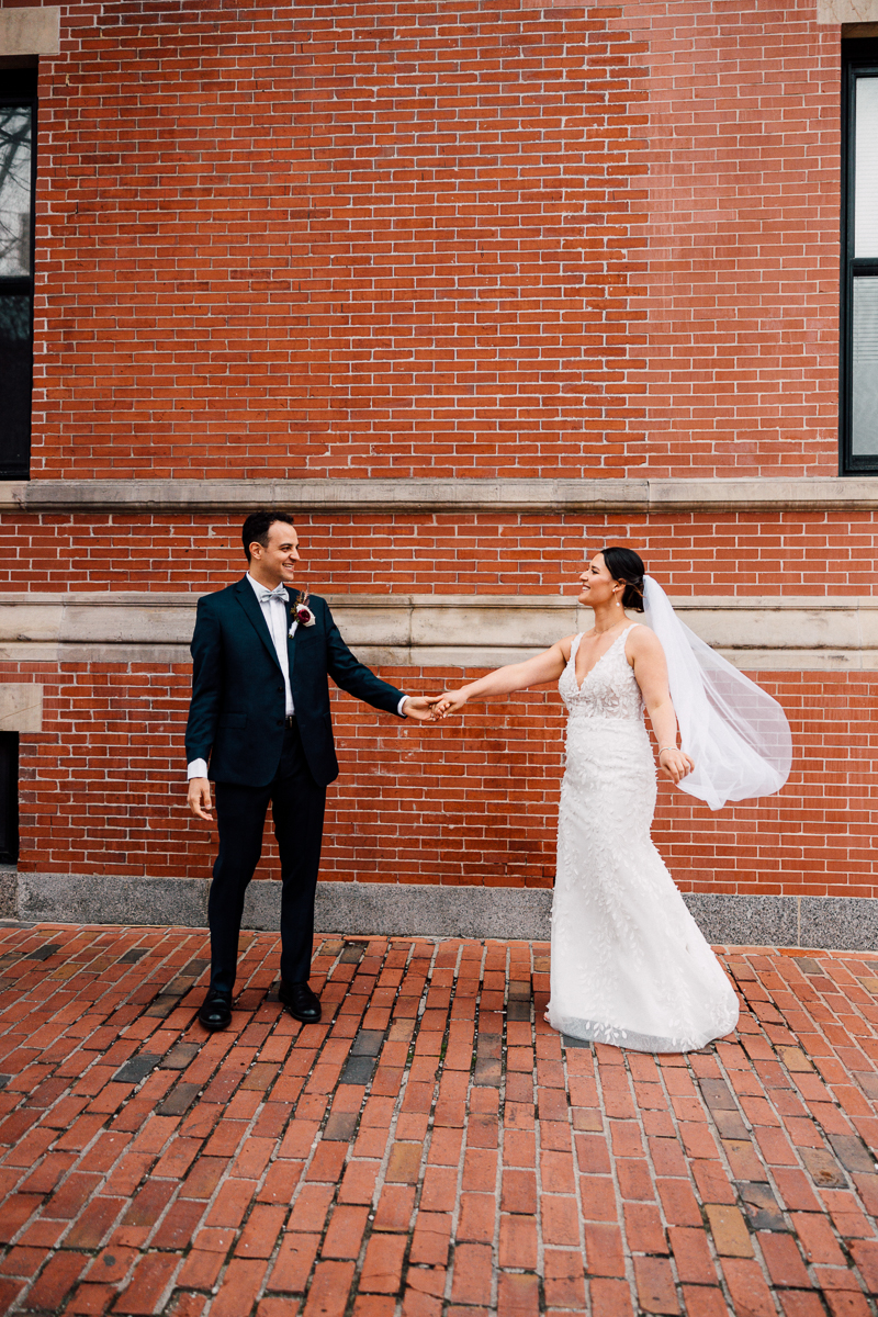 Joyful Wedding Moment::A bride and groom hold hands and smile at each other in front of a brick wall, showcasing a moment of love and connection.