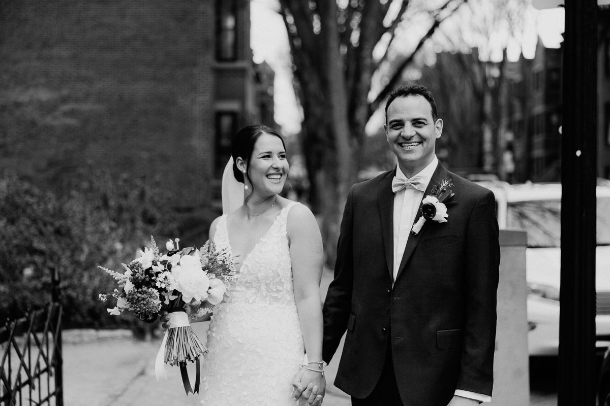 A joyful couple holding hands, with the bride in a lace wedding dress and bouquet, and the groom in a suit, smiling outdoors.
