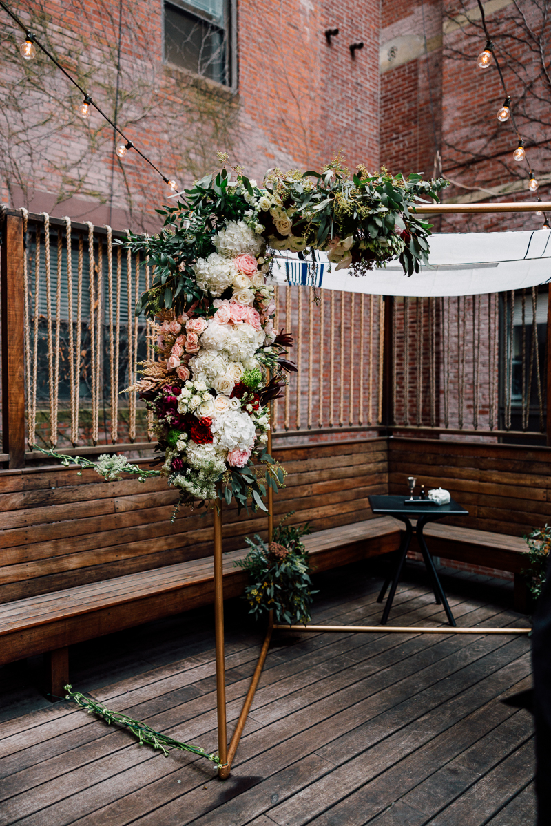 A beautifully arranged floral arch made of various blooms, enhancing a cozy outdoor seating area with wooden benches and string lights.