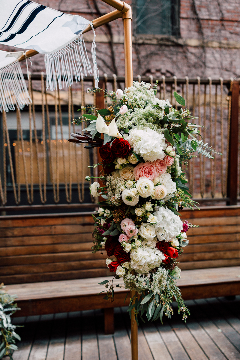 A beautifully arranged floral arch adorned with a mix of roses, hydrangeas, and greenery, alongside a decorative fringed fabric banner, set in a cozy outdoor space.