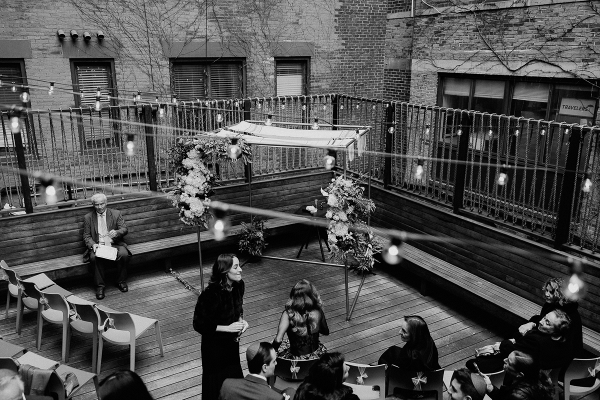 A monochrome view of a beautifully arranged outdoor wedding ceremony featuring floral decorations, seating for guests, and ambient string lights.