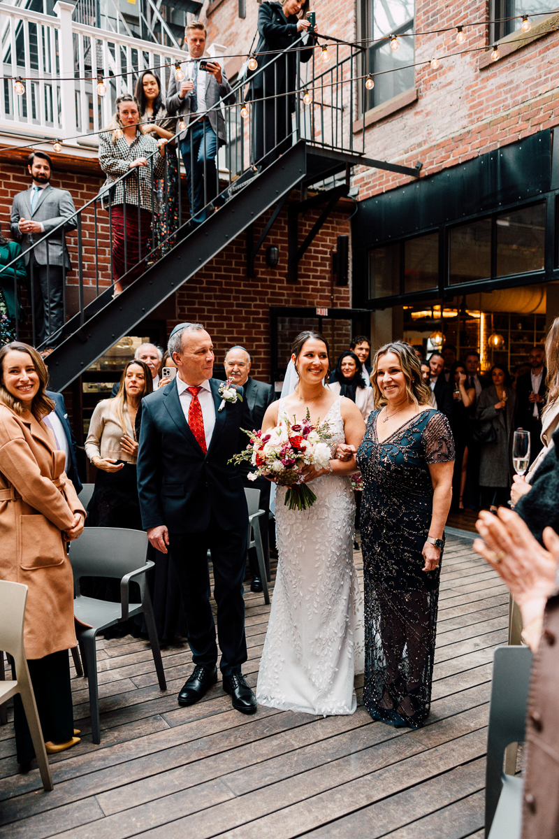 A bride walks down the aisle with her parents, surrounded by guests in a beautifully decorated venue.