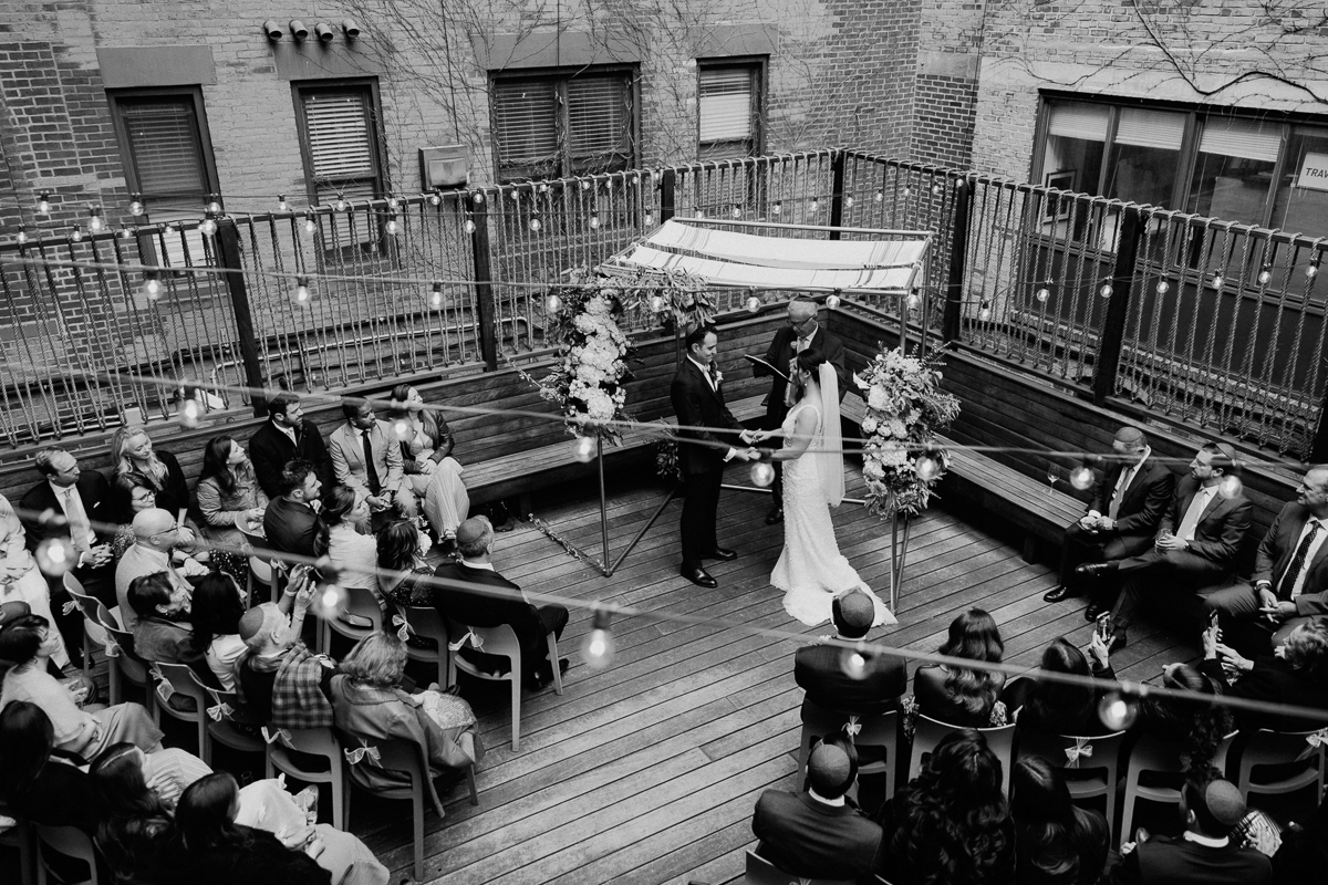 A couple exchanges vows under a floral arch during a rooftop wedding ceremony, surrounded by guests in an intimate setting.