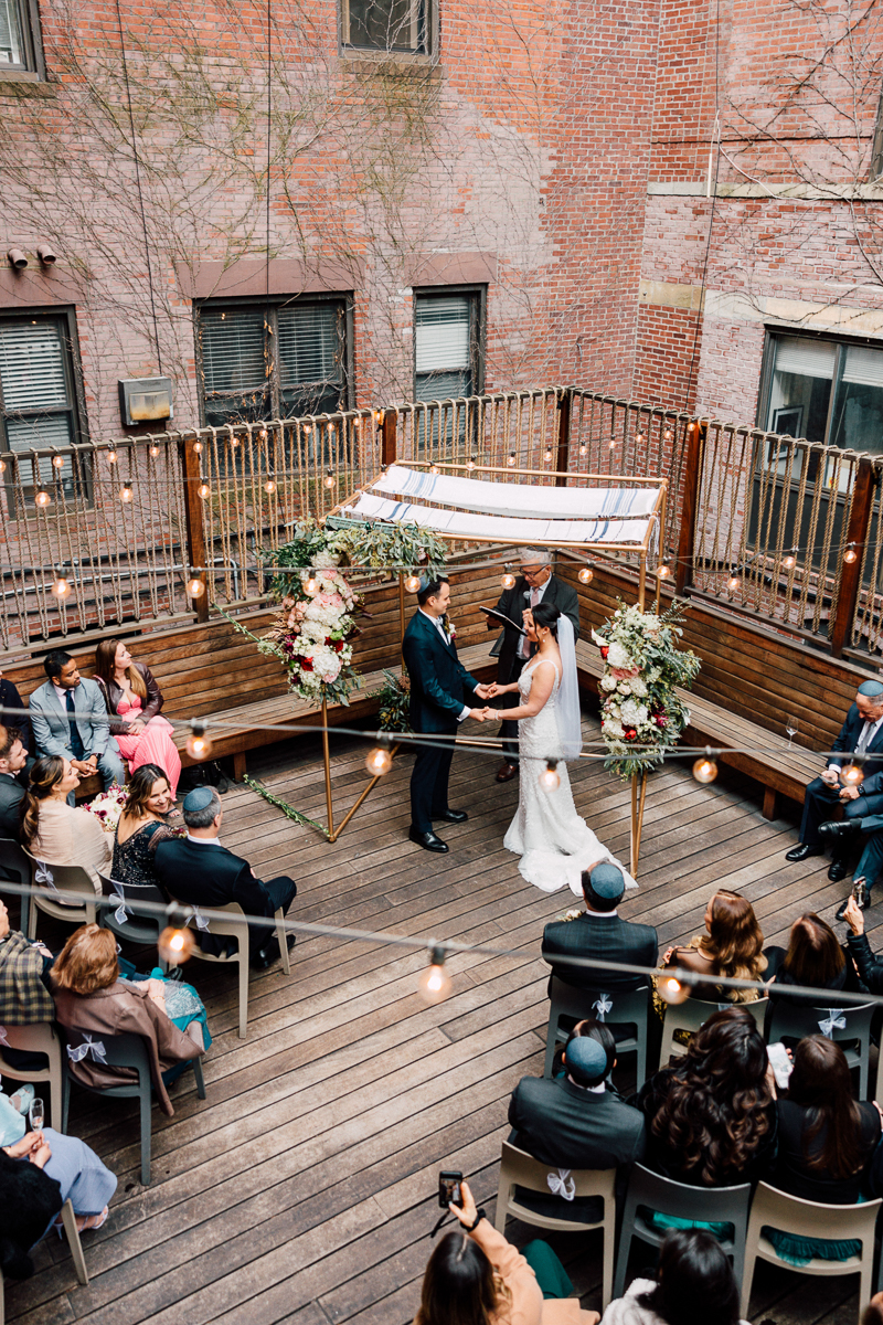 A couple exchanges vows under a beautifully decorated chuppah with floral arrangements, surrounded by seated guests in an intimate outdoor setting.
