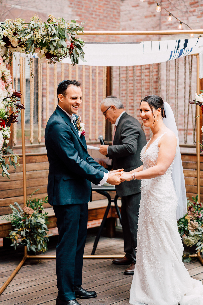 A joyful couple exchanging vows during an intimate wedding ceremony under a floral arch, with a celebrant in the background.