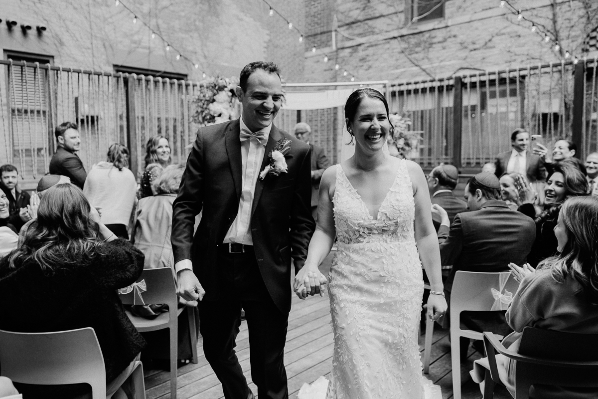 A couple joyfully walks hand in hand down the aisle, surrounded by smiling guests in a beautifully decorated outdoor setting.