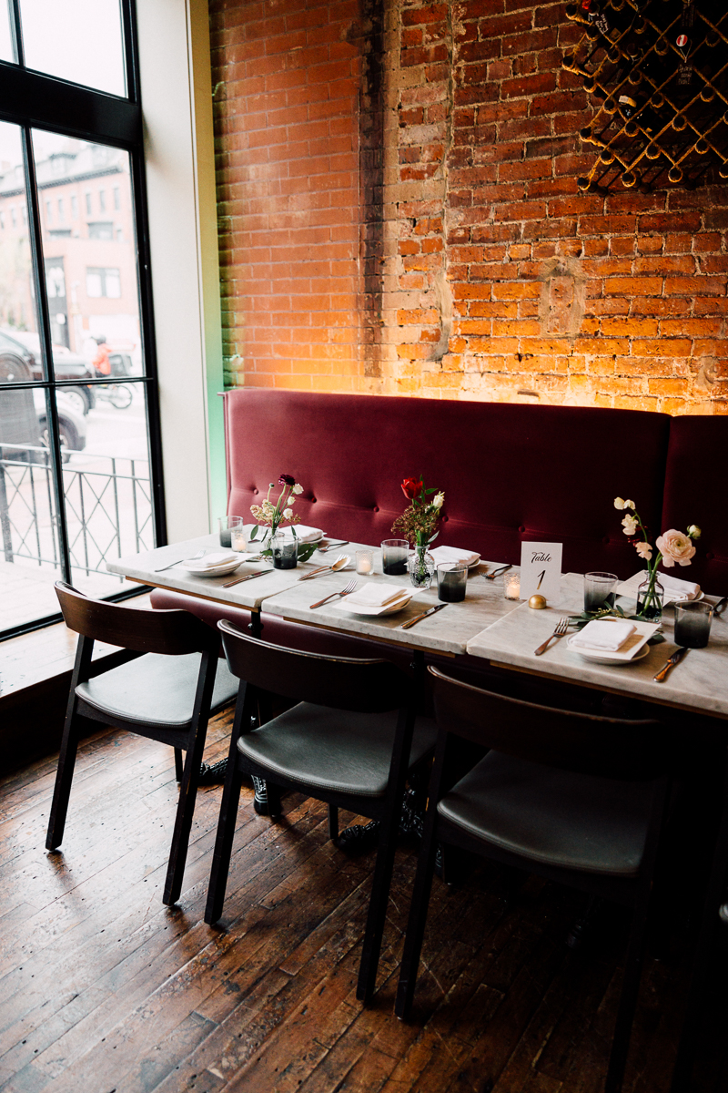 A beautifully arranged dining table featuring a marble top, elegant utensils, and decorative flowers, set against a backdrop of exposed brick and large windows.