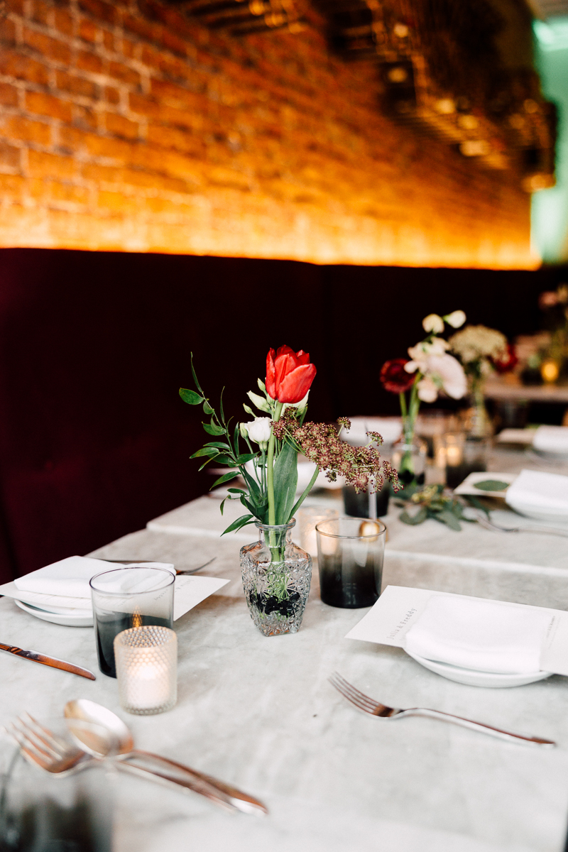 A beautifully arranged dining table featuring a glass vase with red and white flowers, sleek plates, and candlelight ambiance against a brick wall.