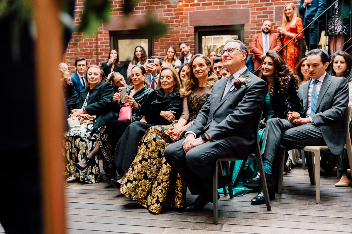 A diverse group of elegantly dressed guests attentively watching a wedding ceremony in a cozy indoor setting.