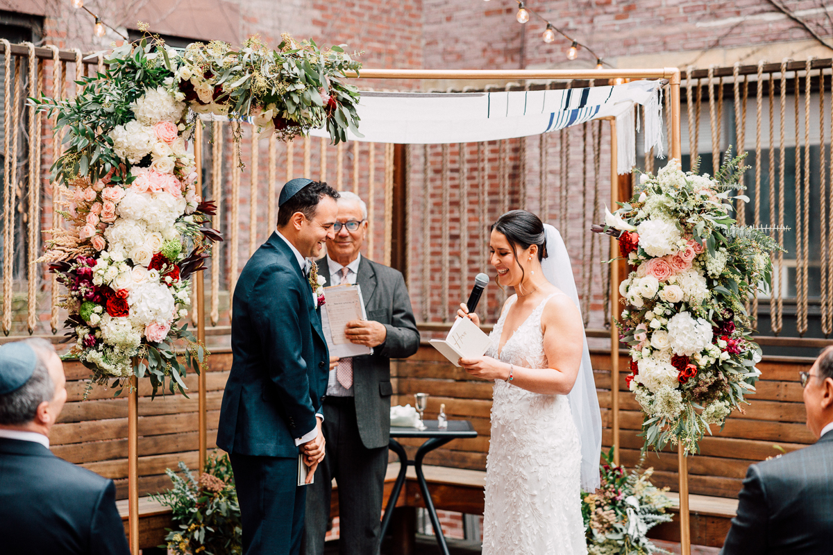 A couple exchanging vows under a floral chuppah during their wedding ceremony, surrounded by guests in a beautiful outdoor setting.