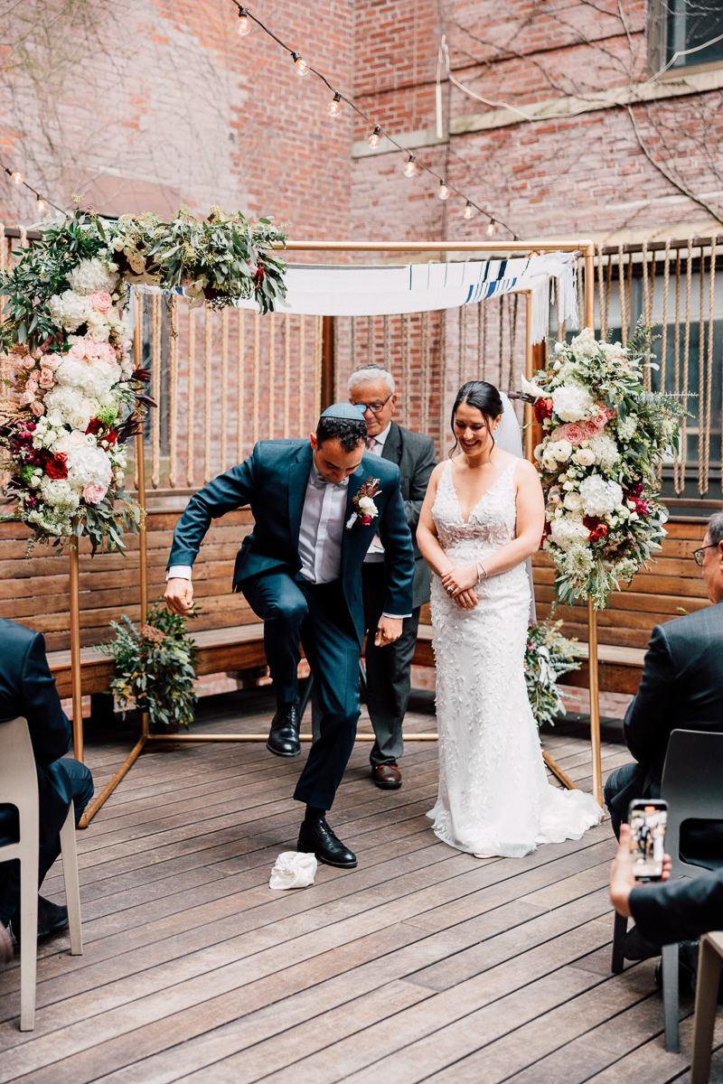 A groom joyfully stomps on a glass during a wedding ceremony under a floral chuppah, while the bride looks on with a smile, surrounded by guests on a wooden platform.