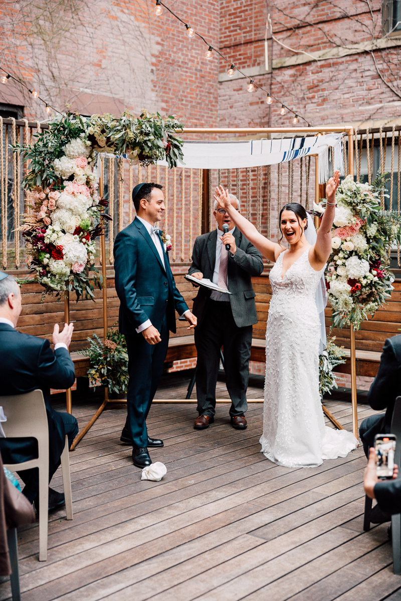 A bride and groom celebrate their wedding under a floral arch, with guests witnessing the joyful moment.