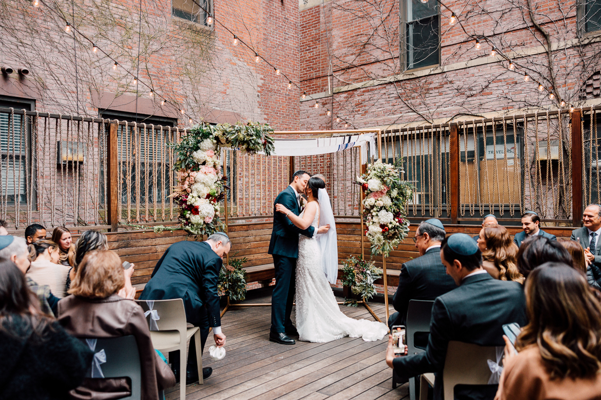 A couple shares a kiss during their wedding ceremony, surrounded by guests in an intimate outdoor space adorned with floral arrangements and string lights.