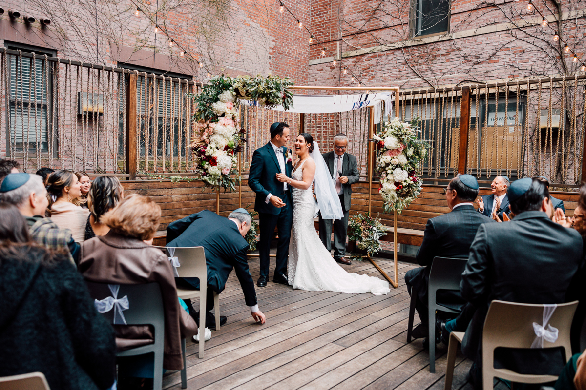 A joyous couple exchanges vows under a floral chuppah during an outdoor wedding ceremony, surrounded by family and friends.