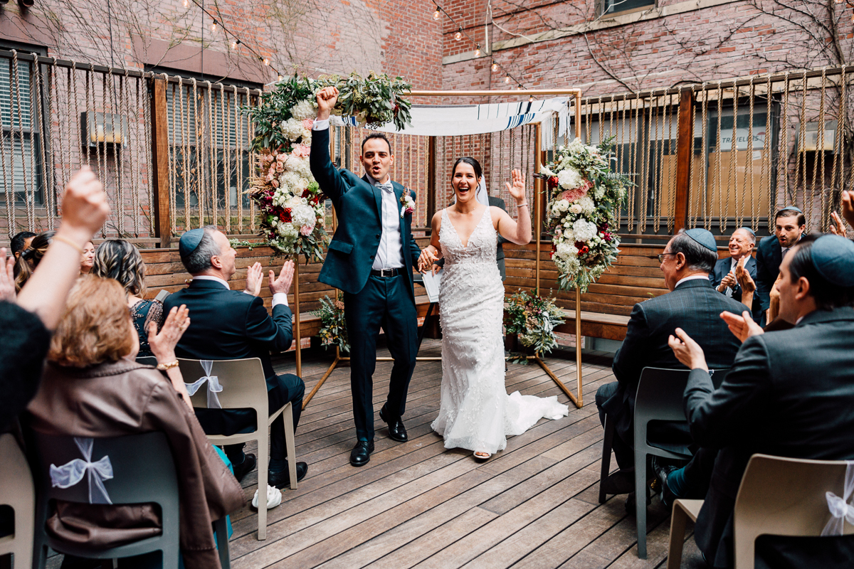 A joyful bride and groom celebrate their marriage as guests applaud in a vibrant courtyard adorned with floral arrangements and a chuppah.