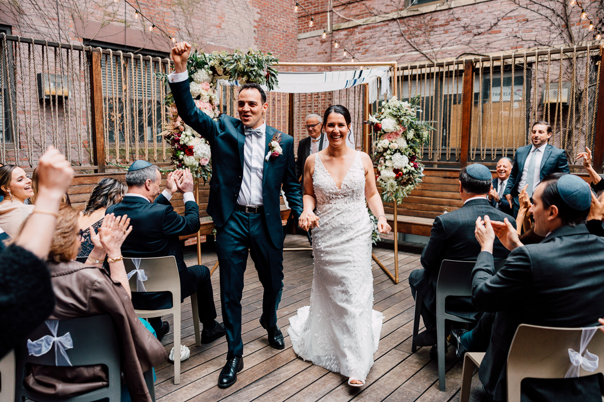 A newly married couple celebrates their union, walking hand in hand and raising their arms in victory, surrounded by applauding guests in an intimate outdoor setting decorated with floral arrangements.
