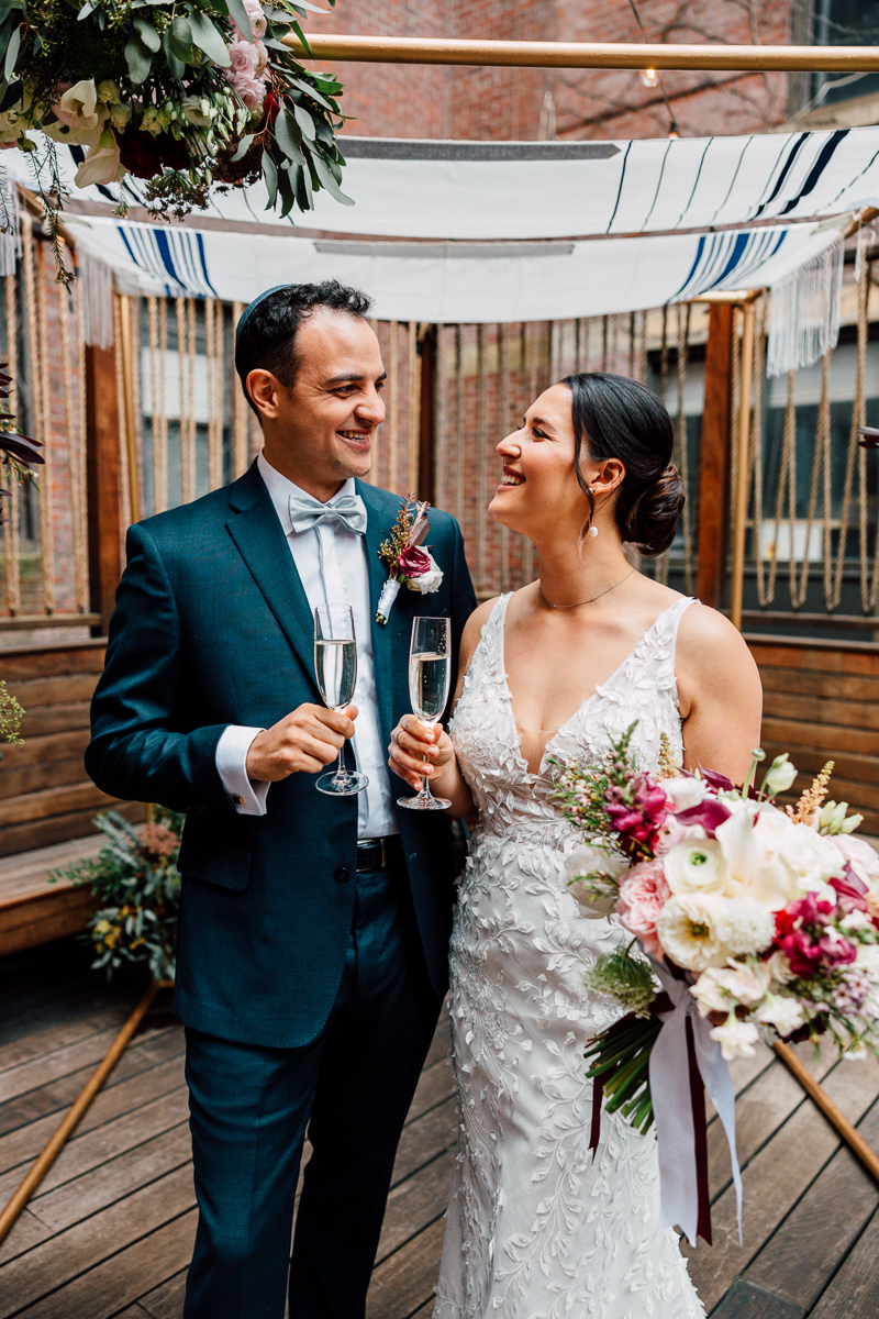 A groom and bride share a joyful moment with champagne glasses in a beautifully decorated outdoor setting, complete with a floral bouquet and festive backdrop.