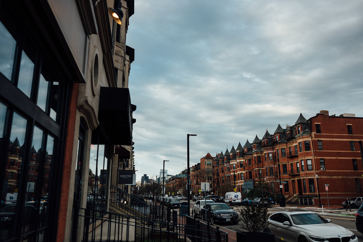 A view of a city street featuring brick buildings, parked cars, and overcast skies, with reflections in storefront windows.