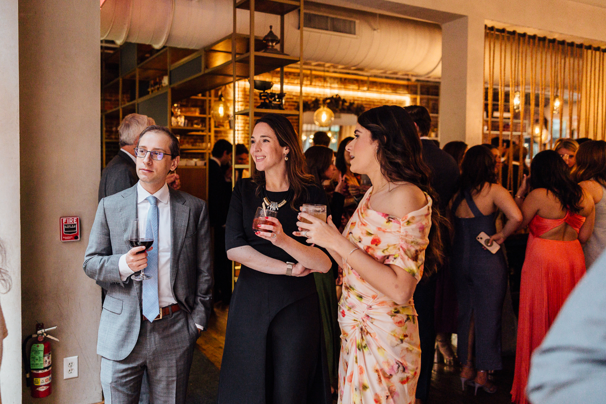 Three well-dressed individuals engaged in conversation at a lively event, with guests mingling in the background.
