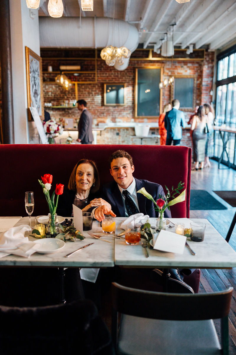 A young man and an elderly woman enjoying a meal together at a beautifully set table with flowers and drinks, in a fashionable restaurant setting.