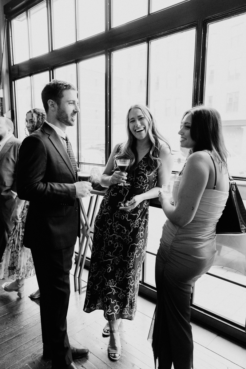 Three adults enjoying drinks and conversation near large windows at a stylish event.