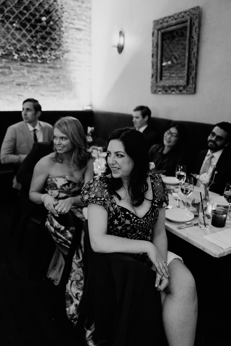 A group of elegantly dressed individuals seated at a dinner table, engaged in conversation, with a warm and intimate atmosphere highlighted by soft lighting and decor.