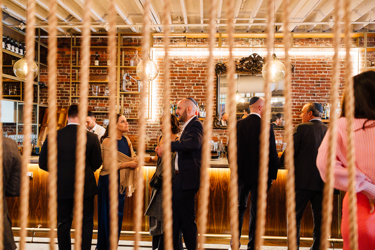 Guests engaged in conversation at a stylish bar with a rustic brick backdrop and hanging lights.
