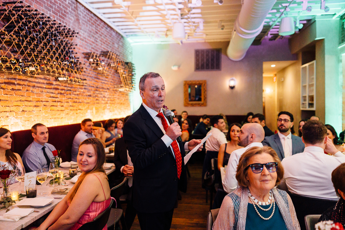 A man in a suit gives a speech while attendees listen attentively at a festive dinner gathering.