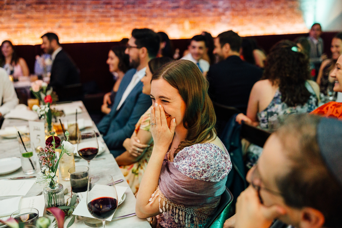 A woman at a dinner party smiles and covers her face in laughter, surrounded by guests enjoying the lively atmosphere.