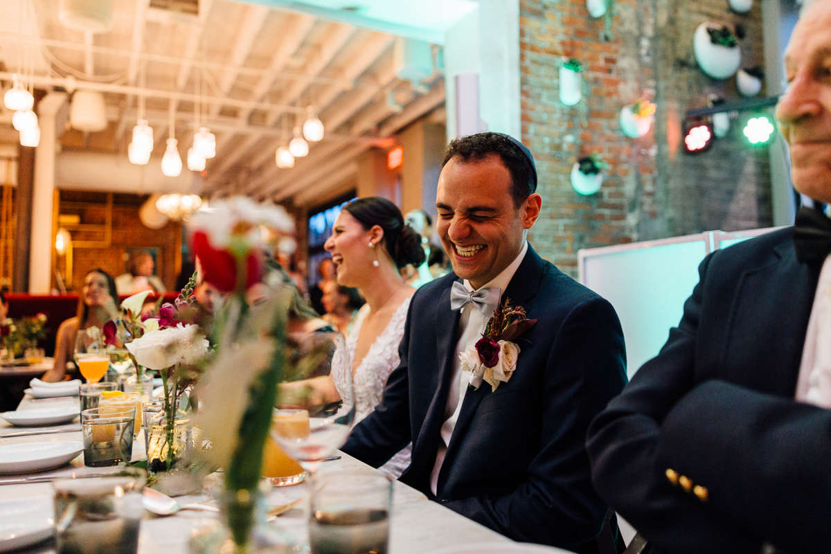 A groom laughing heartily at a wedding reception, surrounded by happy guests and elegant decor featuring flowers and soft lighting.