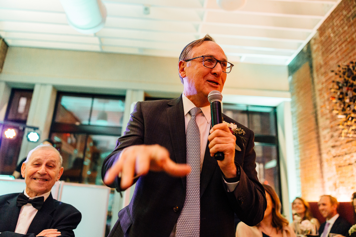 A man in formal attire delivers a speech with a microphone at a wedding reception, while an older man in a tuxedo smiles in the foreground.