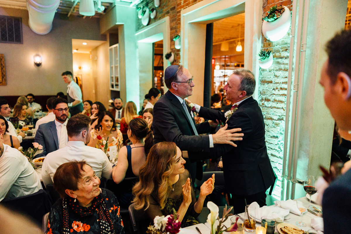 Two men warmly greet each other in a lively restaurant setting filled with guests enjoying a festive atmosphere.