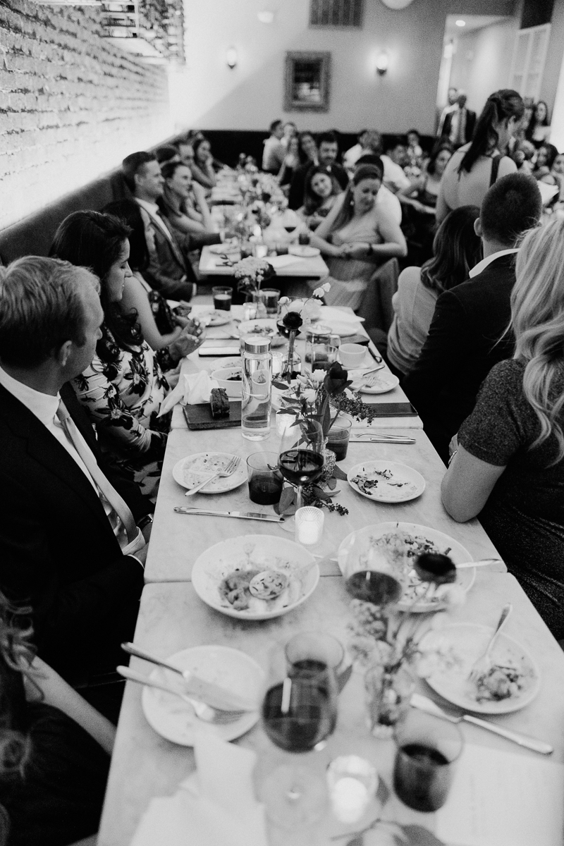 A black and white image of guests seated at a long dining table, enjoying a meal with decorative flowers and various dishes, set in a cozy restaurant atmosphere.