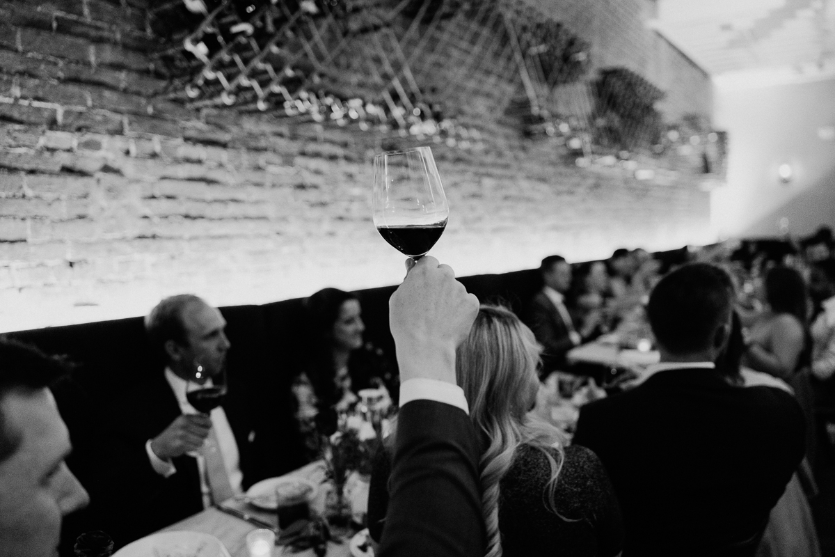 A hand raised with a glass of red wine amidst a festive gathering, featuring guests seated at a beautifully set table in a warmly lit venue.