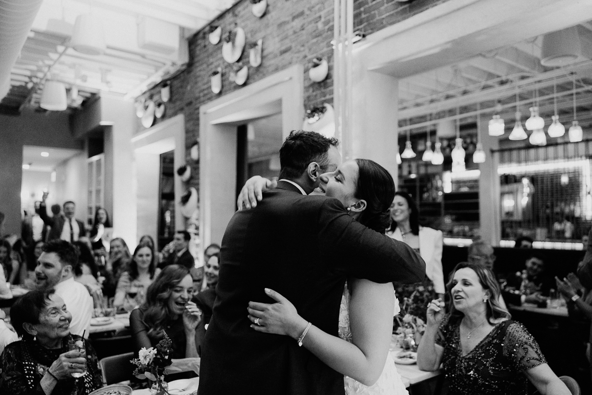 A joyful couple shares a heartfelt embrace during their wedding reception, surrounded by family and friends in a warmly lit venue.