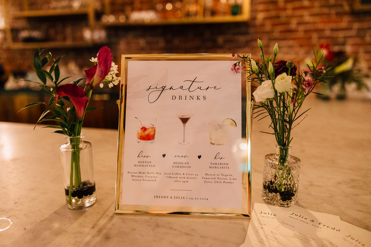 A beautifully framed menu showcasing signature drinks alongside elegant floral arrangements on a marble surface.