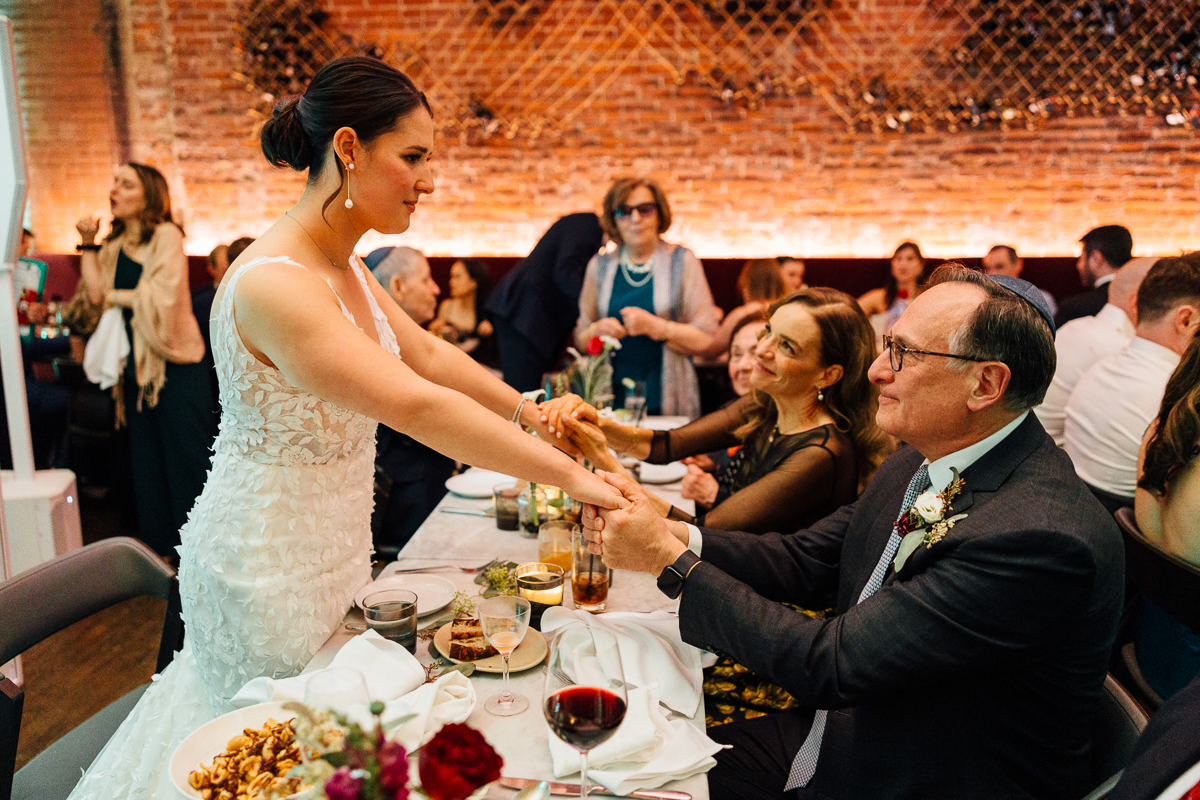 A bride extending her hand while interacting warmly with a seated guest during a wedding reception, surrounded by guests enjoying the event.