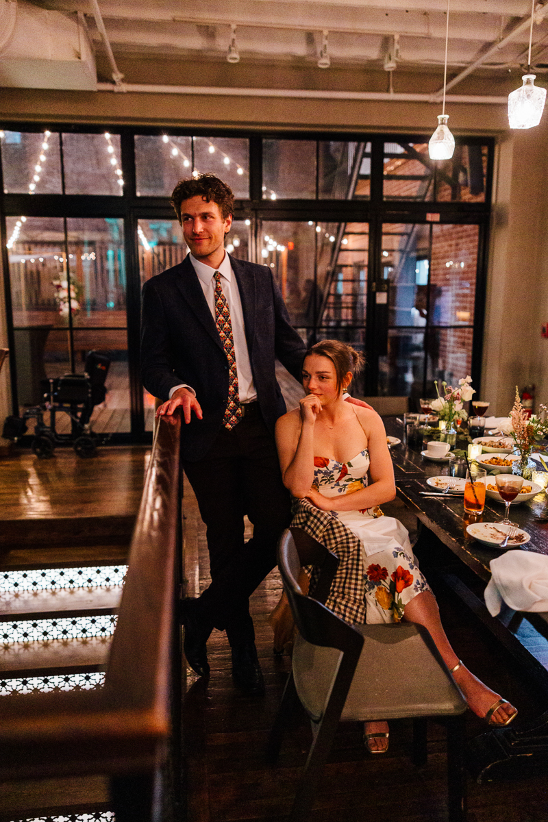 A man in a dark suit and patterned tie stands by a wooden railing, while a woman in a floral dress sits nearby, both in an elegantly decorated restaurant setting.