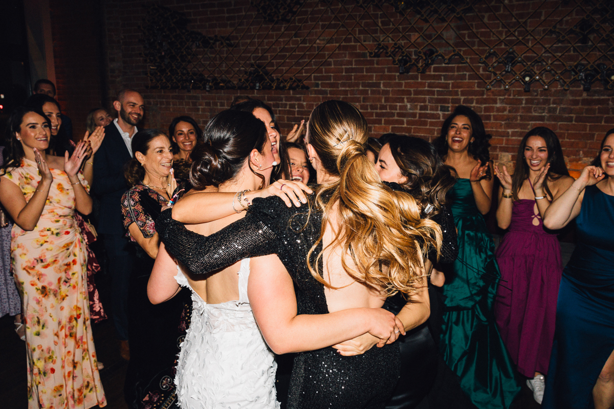 A joyous group of women embrace and celebrate together at a wedding reception, surrounded by smiling guests in elegant attire.