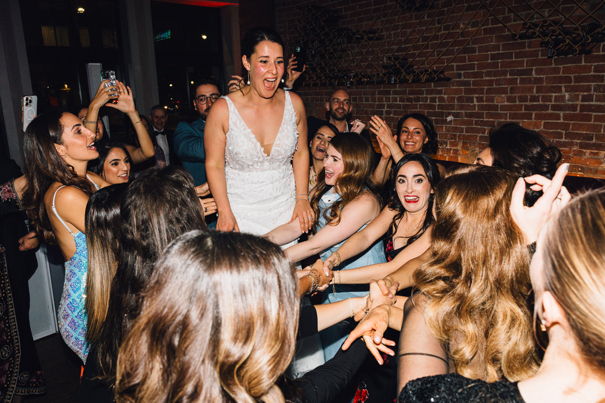 A bride joyfully being lifted by friends during a lively wedding celebration, surrounded by excited guests.