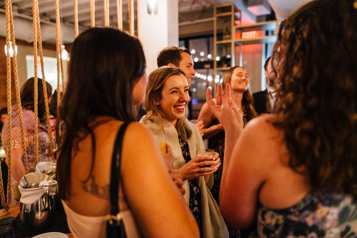 A lively group of people enjoying drinks and engaging in conversation at an event, with one woman smiling brightly in the center.
