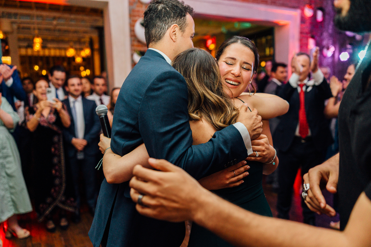 A heartfelt moment of three friends embracing in a joyful celebration, with guests enjoying the lively atmosphere in the background.