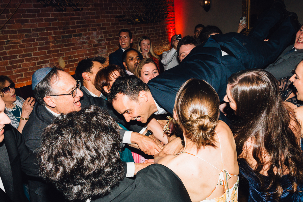 A lively crowd enjoys a festive moment as a man is lifted above them, surrounded by joyful expressions and laughter.