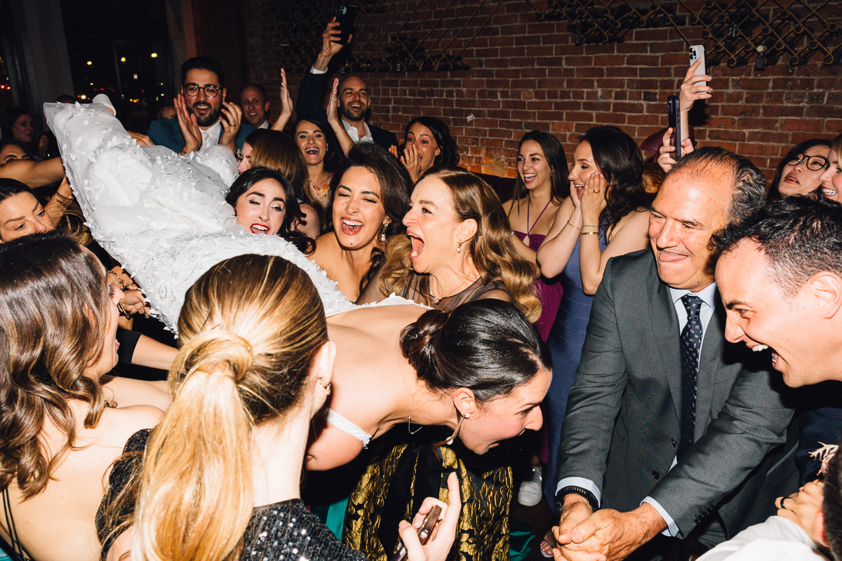 A joyful crowd gathers to celebrate as a bride is lifted in her wedding dress, surrounded by friends and family, all sharing laughter and excitement.