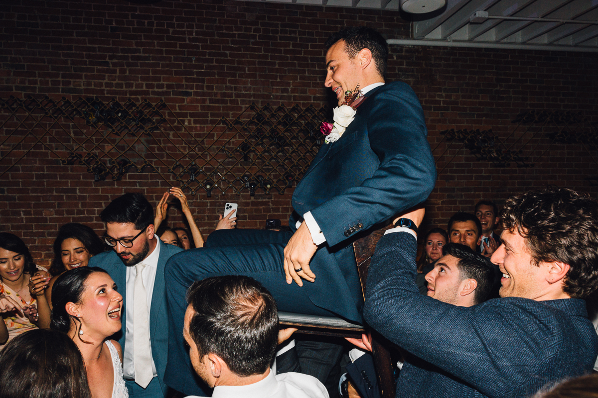 A groom being lifted on a chair by friends during a lively wedding celebration, with guests cheering and enjoying the moment.