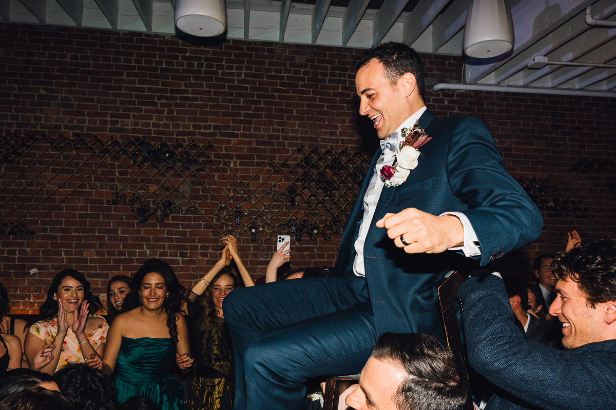 Groom being lifted on a chair amidst a joyful crowd during a wedding reception, with guests clapping and smiling in celebration.