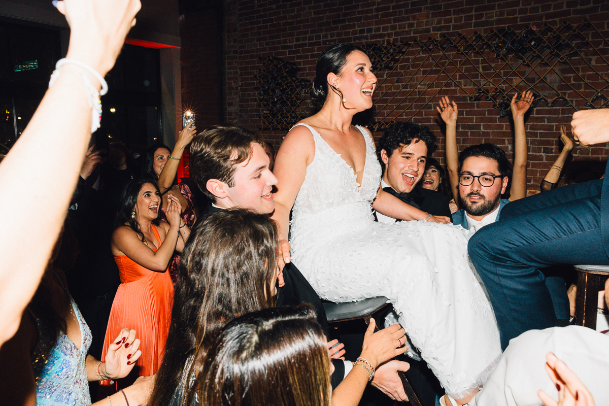 A bride is joyfully lifted by her friends during a lively wedding celebration with guests dancing and cheering in the background.